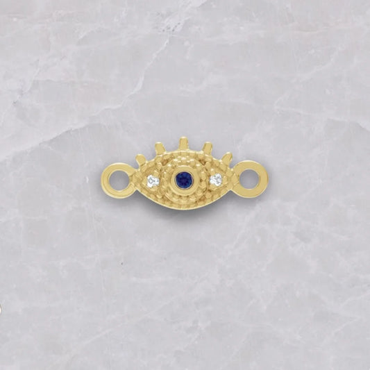 14k gold evil eye connector shown on a white marble background. The connector is an evil eye with loops on the side, 1 natural diamond on each side of the eye, and sapphire as the pupil. 